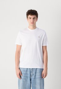 Camiseta de algodón blanca con mangas cortas y cuello redondo. Presenta un pequeño logo en un cuadrado en el pecho. Combinada con unos vaqueros de mezclilla azul claro.