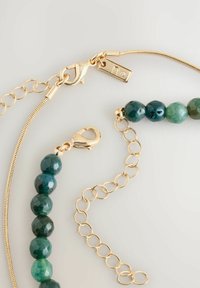 Goldkette und Armband mit grünen, polierten runden Perlen und Karabinerverschluss auf hellgrauem Hintergrund.