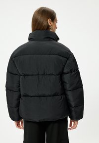 Chaqueta puff negra, con cuello alto, diseño acolchado y textura suave. Vista trasera que muestra una silueta sobredimensionada y detalles en los puños.