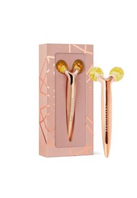 Rouleau facial en or rose avec des embouts en cristal jaune, présenté dans une boîte rose douce ornée de motifs géométriques et du nom de la marque en lettres dorées.