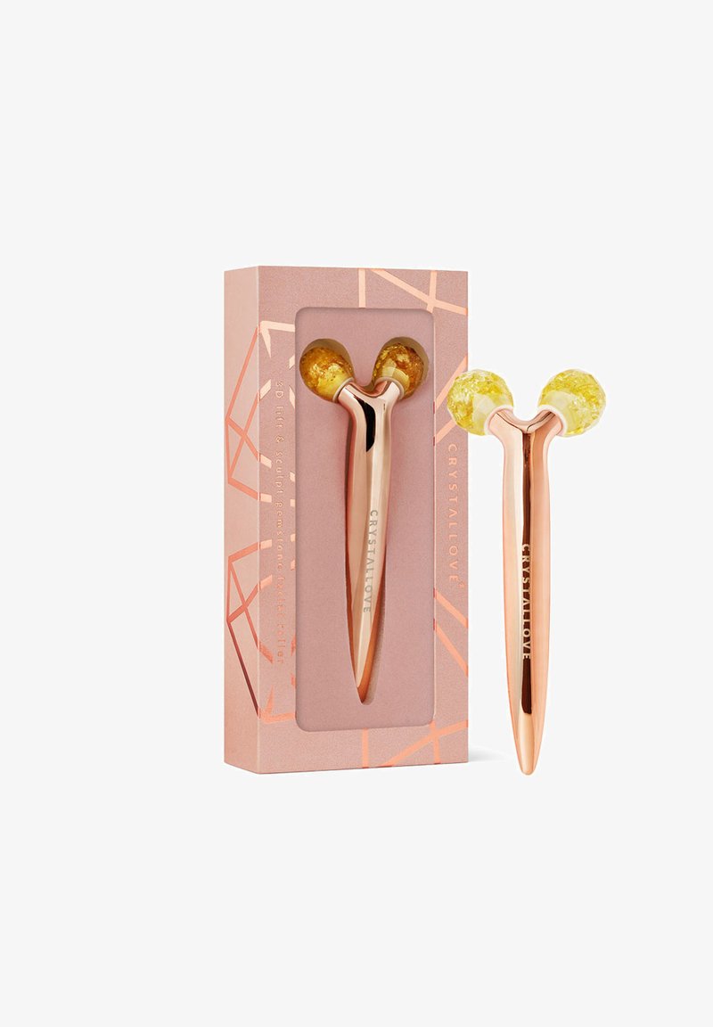 Rouleau facial en or rose avec des embouts en cristal jaune, présenté dans une boîte rose douce ornée de motifs géométriques et du nom de la marque en lettres dorées.