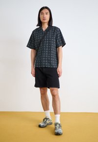 Kortærmet sort skjorte med hvid geometrisk mønster, parret med sorte shorts og grå sneakers. Modellen står på et gult gulv.