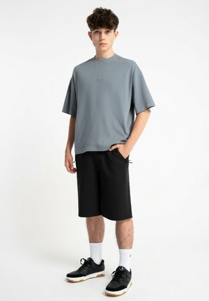 Młody mężczyzna stoi, ubrany w luźny szary t-shirt, czarne szorty do kolan, białe skarpetki i czarne trampki na białym tle.