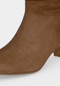 Bottine en daim marron avec un petit talon bloc et un bout pointu arrondi sur fond blanc.