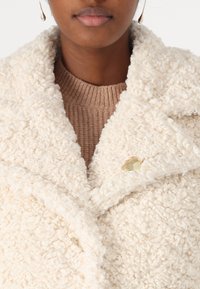 Crème-kleurige, textuurige fuzzy jas met een grote kraag en een gouden accentknop, gelaagd over een bruine geribbelde trui.