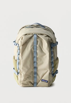 Smėlio spalvos Patagonia kuprinė su mėlynomis rankenomis, akių šoninėmis kišenėmis, viršutine rankena ir priekinės pusės logotipo lopeliu, rodoma vertikalioje padėtyje ant balto fono.