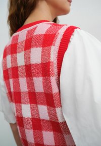 Kobieta w czerwono-różowej kratkowanej dzianinowej kamizelce noszonej na białej bluzce z bufiastymi rękawami, pokazana z górnej części pleców i z boku.