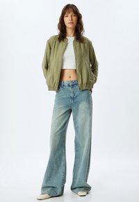 Veste bomber verte olive avec des poignets élastiques, portée sur un crop top blanc, associée à un jean large bleu clair et des chaussures ivoire.
