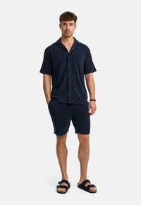 Marineblaues Kurzarm-Hemd mit Knopfleiste, kombiniert mit passenden Shorts. Hergestellt aus strukturiertem Stoff; getragen mit dunklen Sandalen und minimalem Schmuck.