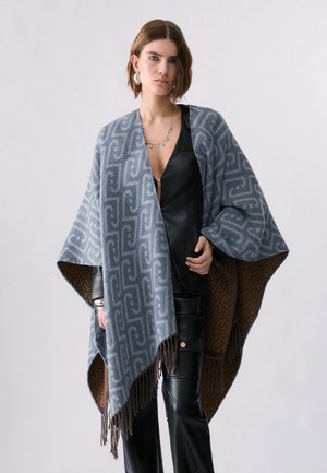 Donna che indossa un mantello con frange blu e marrone a motivi sopra un completo in pelle nero e gioielli d'argento su uno sfondo grigio uniforme.