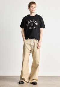 Črna bombažna majica s poudarjenim logotipom "CALVIN KLEIN", v kombinaciji z svetlo bež širokimi hlačami in črnimi čevlji. Preprost, sodoben dizajn.