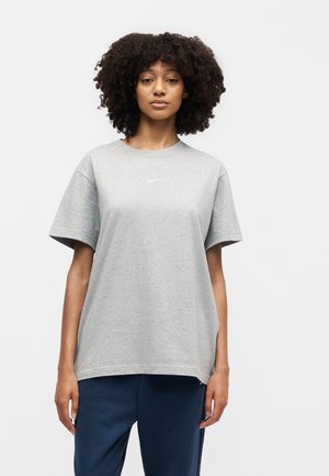 Femme aux cheveux bouclés portant un t-shirt Nike gris ample et un pantalon bleu foncé, debout devant un fond clair uni.
