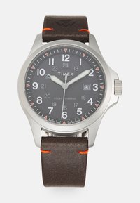 Montre Timex à énergie solaire avec un cadran gris foncé, des chiffres blancs, un boîtier en acier inoxydable et un bracelet en cuir marron avec des coutures orange.