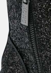 Bottine noire à sequins avec une fermeture éclair lisse et une étiquette métallique embossée du mot "KORS". Présente une surface texturée et réfléchissante.