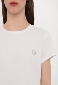 Bílý tričko s krátkým rukávem vyrobené z hladkého materiálu, s malým šedým kruhovým logem s nápisem "AX" na levé části hrudi.