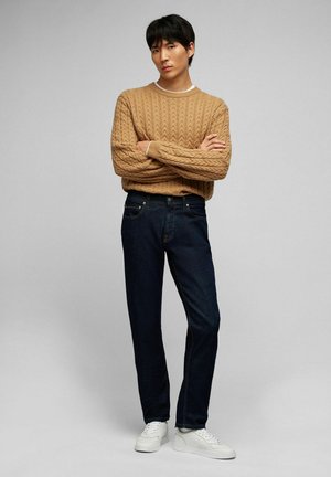 Beige gestrickter Pullover mit Kabeldesign, kombiniert mit dunkelblauen Jeans und weißen Sneakers. Das Model steht mit verschränkten Armen vor neutralem Hintergrund.