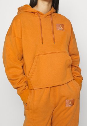 Person wearing matching orange hoodie and sweatpants with front pockets and small branded leather patches on chest and thigh.