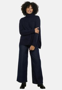 Jūras zila, brīvā piegriezuma turtlenecks, ar ribinātām piedurknēm un apakšmalu, apvienots ar plaša piegriezuma tumšajiem džinsiem un melniem potīšu zābakiem.