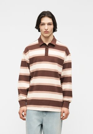 Mann mit mittellangen dunklen Haaren, der ein braunes, beige- und cremefarben gestreiftes Langarm-Poloshirt und hellblaue Jeans vor einem weißen Hintergrund trägt.