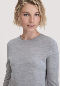 Blonde vrouw met kort golvend haar in een lichtgrijze lange mouw top, die recht vooruit kijkt met een neutrale achtergrond.