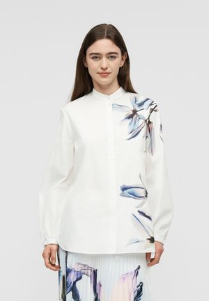 Mladá žena s dlhými hnedými vlasmi nosí bielu košeľu na gombíky s modrými a fialovými kvetinovými vzormi na ramene a boku.