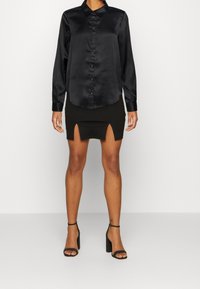Camicia nera di raso a maniche lunghe con colletto e chiusura a bottoni, abbinata a una mini gonna nera con due spacchi frontali. Sandali neri con tacco alto.