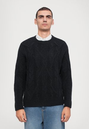 Svart strikket genser med teksturert kabelmønster, rund hals og ribbede mansjetter. Kombinert med lyseblå denimjeans.