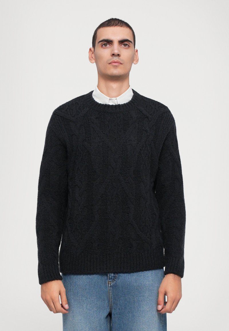 Black knitted sweater with a textured cable pattern, crew neck, and ribbed cuffs. Paired with light blue denim jeans.
