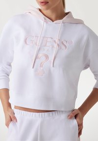 Witte cropped hoodie van zacht materiaal, met een lichtroze capuchon en geborduurd "GUESS" logo met vraagteken detail.