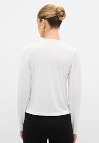 Witte langwerpige top met lange mouwen, aangedreven door een gladde textuur en subtiele schouderdetails. De achterkant heeft een ronde halslijn en een cropped zoom.
