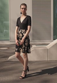 Robe noire à imprimé floral avec un design à enroulement, manches courtes, jupe mi-longue et escarpins noirs. Le modèle tient une petite pochette.