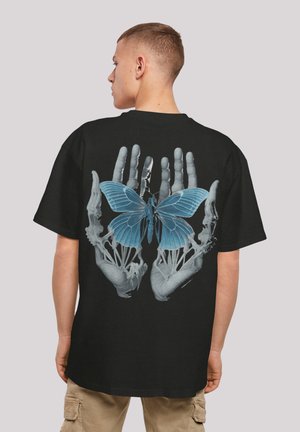 Schwarzes T-Shirt mit einer detaillierten Grafik von Skelethänden, die einen blauen Schmetterling halten, was einen Kontrast aus Farben und Texturen erzeugt.
