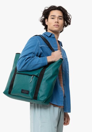 Green tote bag with black accents, made from durable material. Features a front zip pocket and is worn over the shoulder.