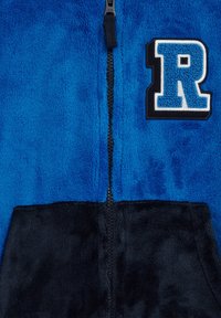 Blå fleece zip-up hoodie med en mjuk textur, svart nederdel och ett blått och vitt bokstavs-patch med "R" på bröstet.