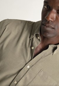 Camisa verde oliva con cuello abotonado, un bolsillo en el pecho y botones de color claro; la tela presenta una textura sutil.