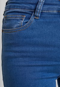 Vaqueros de mezclilla azul con costuras visibles, un pequeño bolsillo frontal y una textura suave, capturando el detalle en primer plano de la tela.