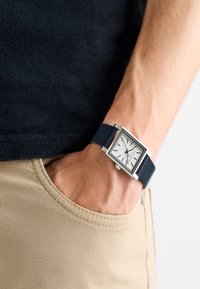 Kvadratisk silverfärgad klocka med vit urtavla, marinblå läderrem, silverfärgade timmarkörer och blå sekundvisare, som bärs på en persons handled.