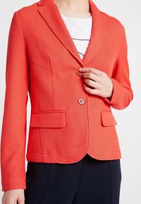 Blazer texturé rouge avec un col cranté, deux poches avant et deux boutons. Porté par-dessus un haut blanc avec des lignes horizontales noires.