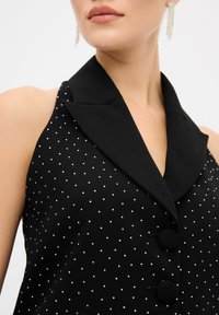 Schwarzes, ärmelloses Oberteil mit einem geschlitzten Kragen, verziert mit silbernen Punkten und zwei runden, schwarzen Knöpfen. Glatte Stofftextur.