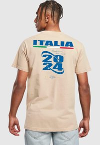 Beige pamut póló, melyen kék grafika található "2024" és "ITALIA" felirattal, zöld és piros kiegészítőkkel, hátulról nézve.