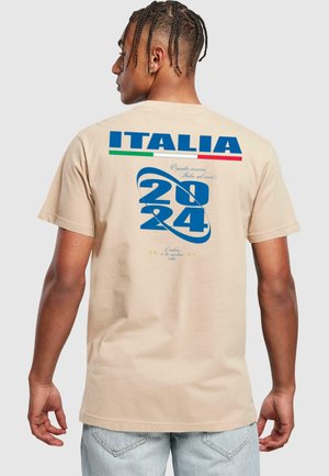 Beige pamut póló, melyen kék grafika található "2024" és "ITALIA" felirattal, zöld és piros kiegészítőkkel, hátulról nézve.