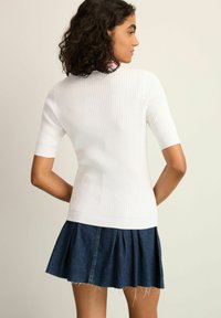 Weißes kurzärmeliges Top mit Rippenstruktur und figurbetontem Schnitt, kombiniert mit einem plissierten Jeansrock mit fransigem Saum. Sichtbare Textur- und Formdetails.