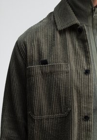 Kordhemdjacke in Olivgrün, mit vertikaler gerippter Textur, einer knopfverschlossenen Tasche und einem Kragen mit Reißverschluss.