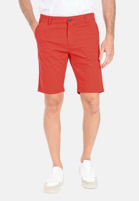 Pantaloni corti di cotone rossi con un bottone blu, tasche laterali e taglio dritto, abbinati a sneakers bianche su uno sfondo neutro.