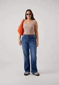 Mulher vestindo uma blusa de alças listrada, jeans azuis, sandálias, bolsa laranja e óculos de sol, posando contra um fundo claro e simples.