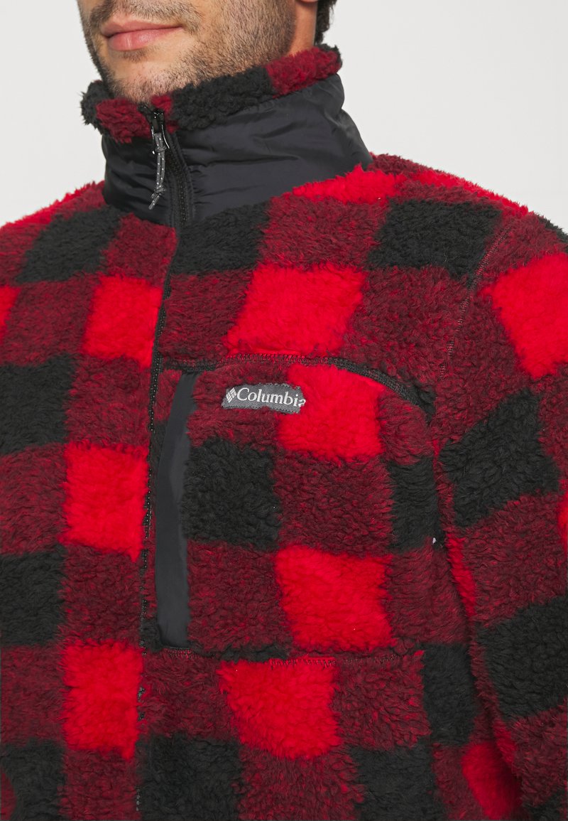 Veste en polaire à carreaux rouges et noirs avec un col haut, fermeture éclair, matériau texturé et poche poitrine arborant un logo Columbia.