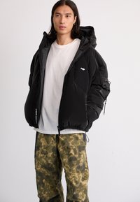 Black puffer jacket with a hood, featuring a front zipper, side pockets, and a logo. Paired with camo-patterned green pants.