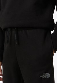 Sudadera negra con cintura ajustable mediante cordón y bolsillos delanteros, confeccionada en tejido texturizado. Presenta el logotipo de la marca en la parte inferior izquierda.