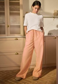 Vrouw die in een keuken staat, gekleed in een wit T-shirt met korte mouwen, losse perzikkleurige gestreepte broek en beige schoenen, met haar handen in haar zakken.