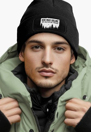 Black ribbed beanie with a white patch featuring text and forest graphic. Layered with a light green waterproof jacket and black inner lining.